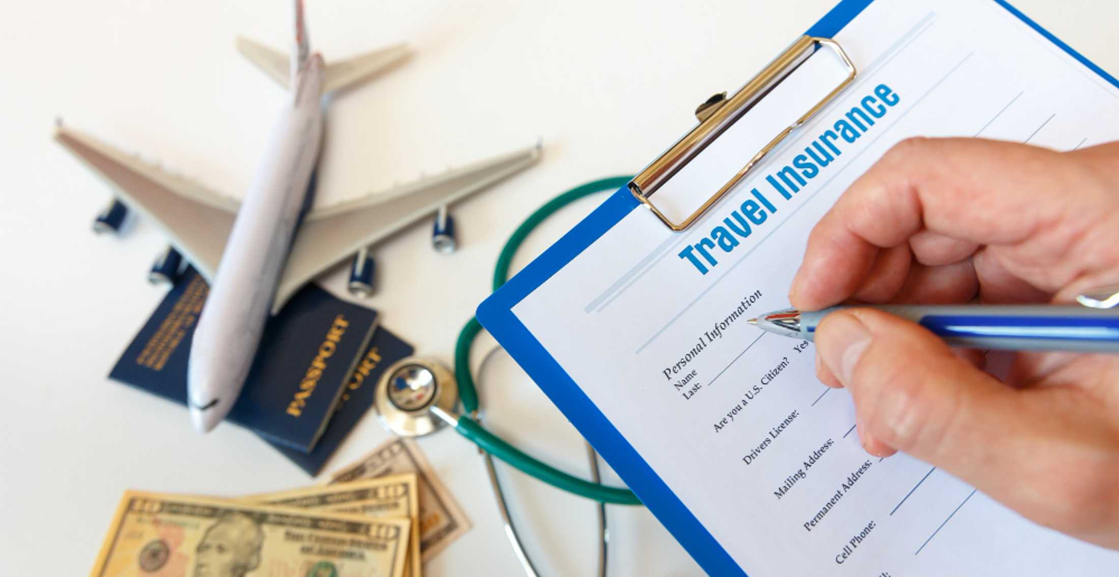 Рука заполняет бланк travel insurance рядом с моделью самолёта, паспортами, стетоскопом и деньгами