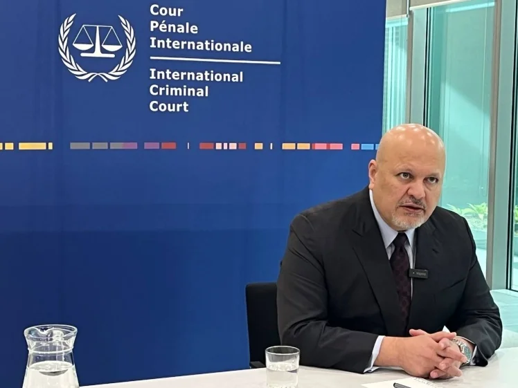 Чоловік у чорному костюмі сидить за столом перед емблемою Міжнародного кримінального суду