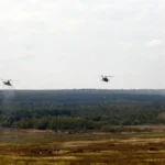 Три гелікоптери летять над лісистим пагорбом і полем під хмарним небом