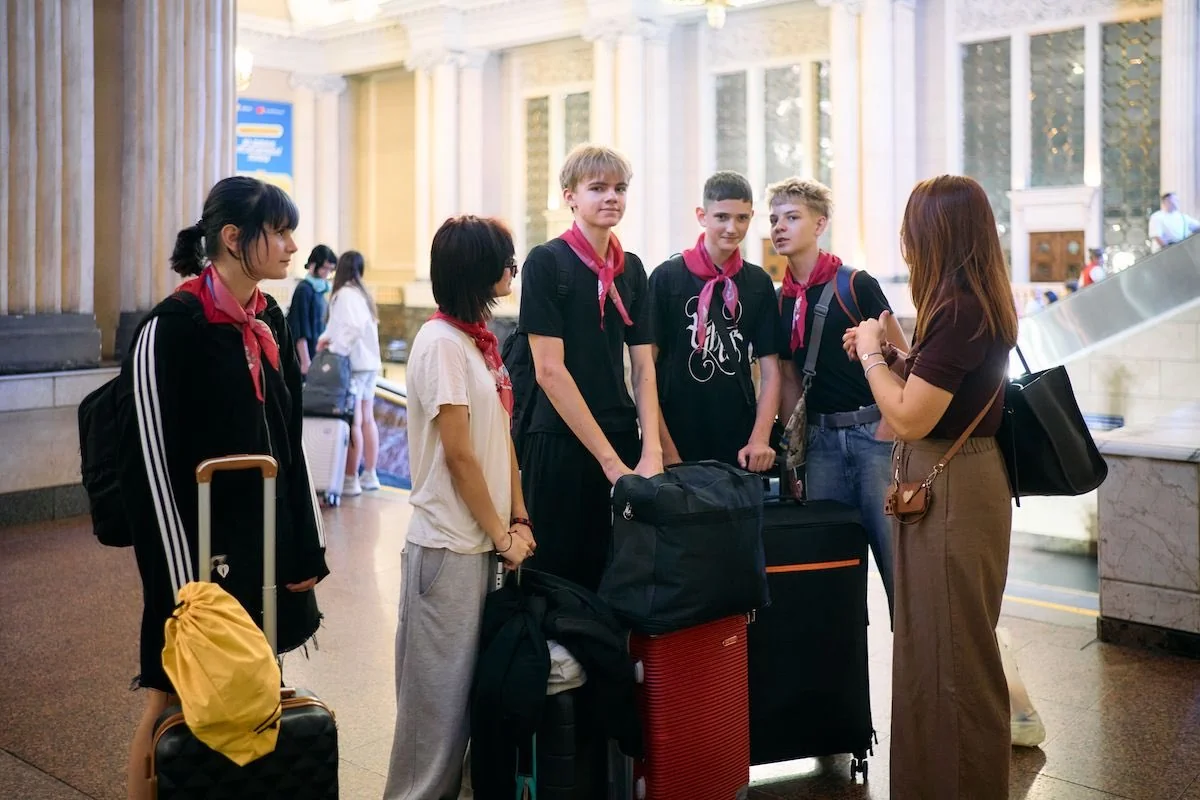 Група підлітків з червоними хустками та валізами спілкується з жінкою у залі