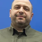 Лисий чоловік з бородою в оливковій сорочці на синьому фоні